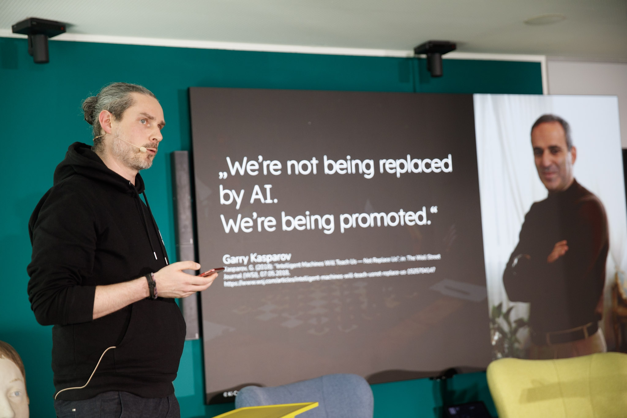Christian Kleemann steht auf einer Bühne und gibt eine KI-Schulung und Weiterbildung. Er zeigt auf eine Leinwand mit einem Zitat zu KI von Garry Kasparov auf dem steht: We're not being replaced by AI. We're being promoted.